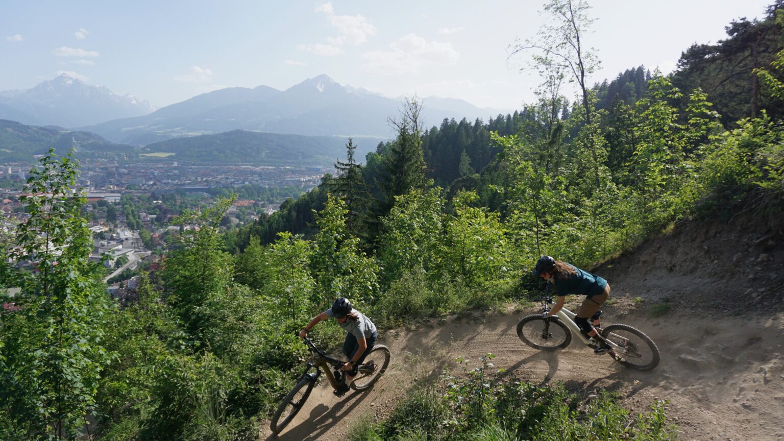 Zwei Radfahrer fahren mit ihren Mountainbikes auf dem Hofwaldtrail, einem gewundenen Feldweg durch eine bewaldete Hügellandschaft, mit einer Stadt und Bergen im Hintergrund.