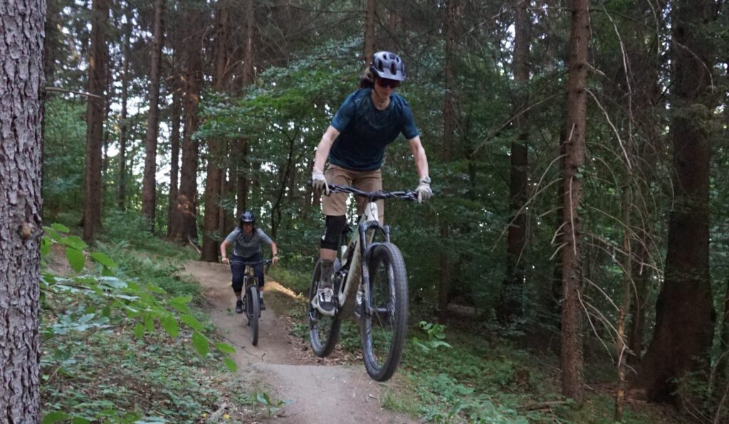Zwei Personen fahren mit dem Mountainbike auf dem Hofwaldtrail in einem Wald; der erste Fahrer fängt nach einem Sprung Luft, während der zweite dicht hinter ihm auf dem unbefestigten Weg folgt.