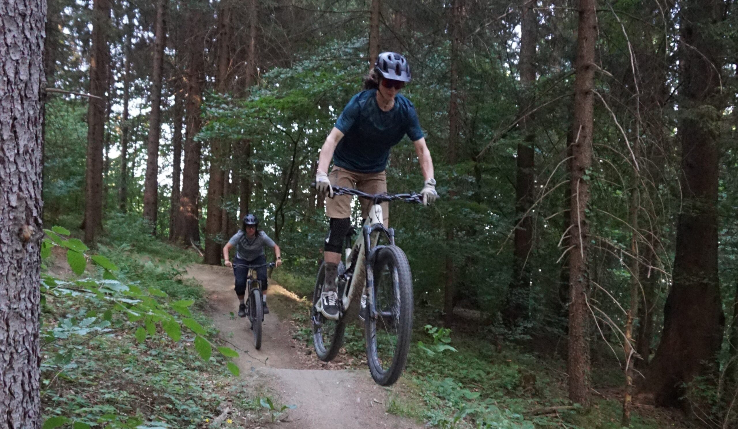 Zwei Personen fahren mit dem Mountainbike auf dem Hofwaldtrail in einem Wald; der erste Fahrer fängt nach einem Sprung Luft, während der zweite dicht hinter ihm auf dem unbefestigten Weg folgt.