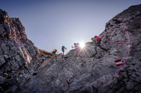 Vier Wanderer mit Rucksäcken steigen im Sonnenlicht einen felsigen Berghang hinauf; auf den Felsen sind rot-weiße Wegmarkierungen zu sehen.