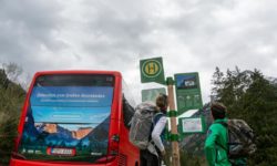 Zwei Wanderer mit Rucksäcken stehen unter einem bewölkten Himmel neben einem Bushaltestellenschild und einem roten Bus, bereit für Wanderabenteuer im Naturpark Karwendel, mit Bäumen und Bergen im Hintergrund.