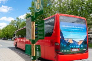 Ein roter Bergbus parkt an einem sonnigen Tag an einem Bushaltestellenschild, mit Bäumen und blauem Himmel im Hintergrund.