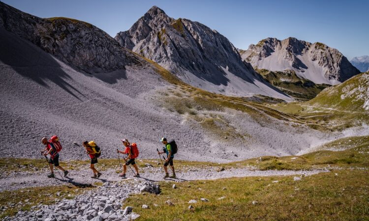 Vier Wanderer mit Rucksäcken wandern auf einem felsigen Bergpfad im Naturpark Karwendel, umgeben von schroffen Gipfeln und grasbewachsenen Hängen unter einem klaren blauen Himmel – perfekt für Wander- und Bergsteigabenteuer.