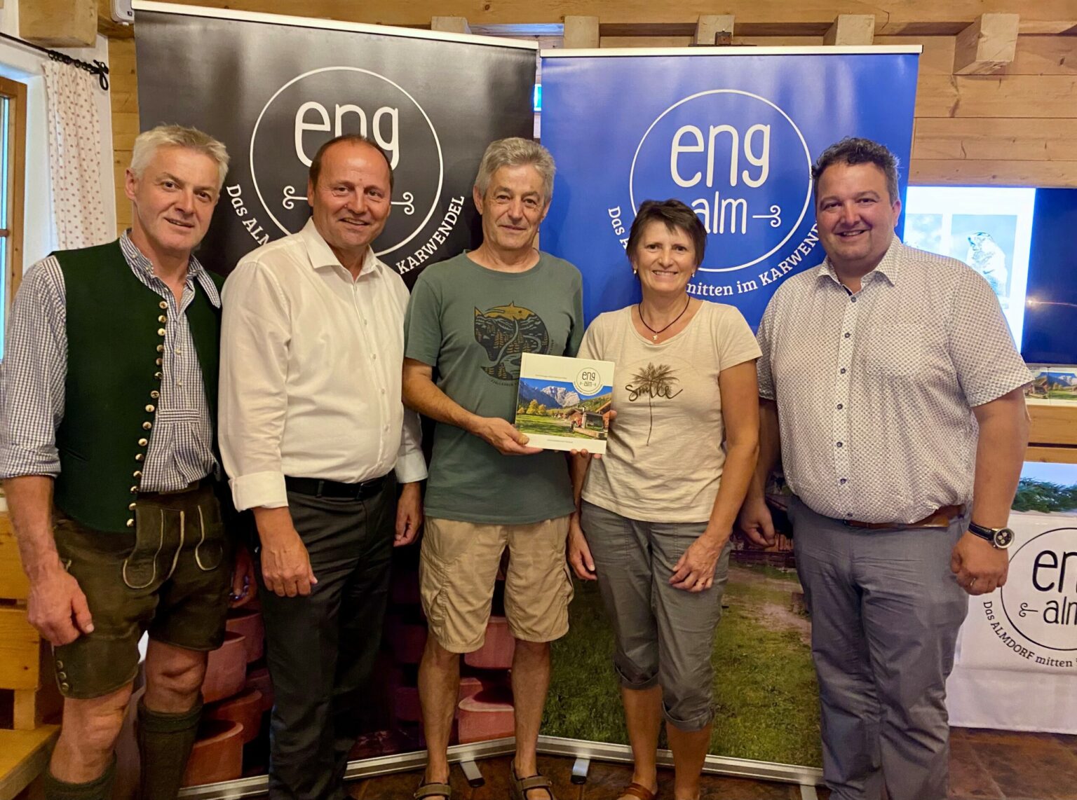 Fünf Personen posieren in einem Innenraum vor "eng alm"-Bannern; die Person in der Mitte hält eine Broschüre mit einem Landschaftsbild, das die Eröffnung Außenbereich des Naturparkhauses im Naturpark Karwendel Hinterriß markiert.