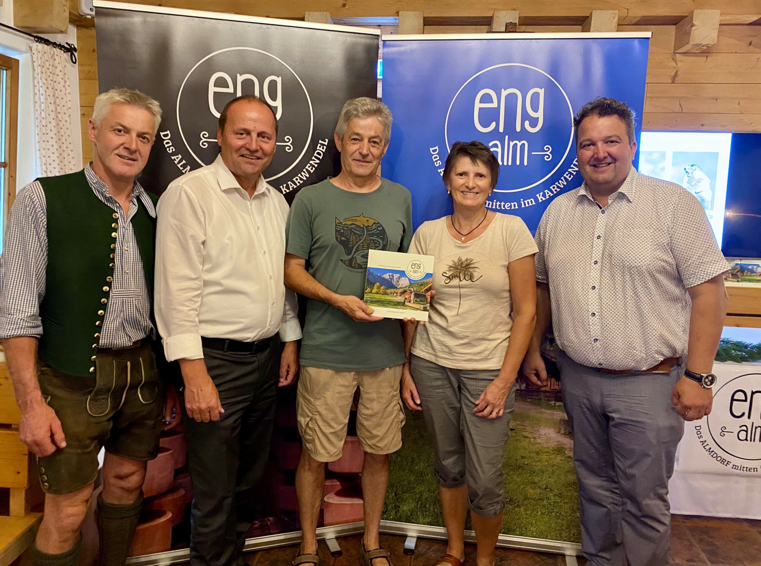 Fünf Personen posieren in einem Innenraum vor "eng alm"-Bannern; die Person in der Mitte hält eine Broschüre mit einem Landschaftsbild, das die Eröffnung Außenbereich des Naturparkhauses im Naturpark Karwendel Hinterriß markiert.