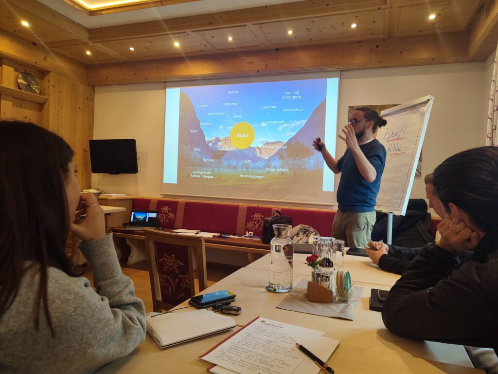 Ein Mann hält einen Vortrag in einem Konferenzraum mit einer Projektionsfläche, auf der ein Diagramm von Mobilität Rißtal 2.0 zu sehen ist, während die Teilnehmer zuhören und sich Notizen machen.