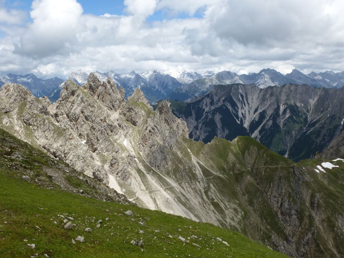 Zackige Berggipfel wie die Erlspitze Bertour und die Reitherspitze erheben sich mit Schneeflecken und grünen Hängen unter einem teilweise bewölkten Himmel, mit fernen schneebedeckten Bergen im Hintergrund.