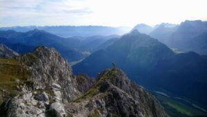 Eine Person steht auf einem felsigen Bergrücken am Mittenwalder Höhenweg Klettersteig und überblickt bei klarem Himmel ein Tal und ferne Bergketten.