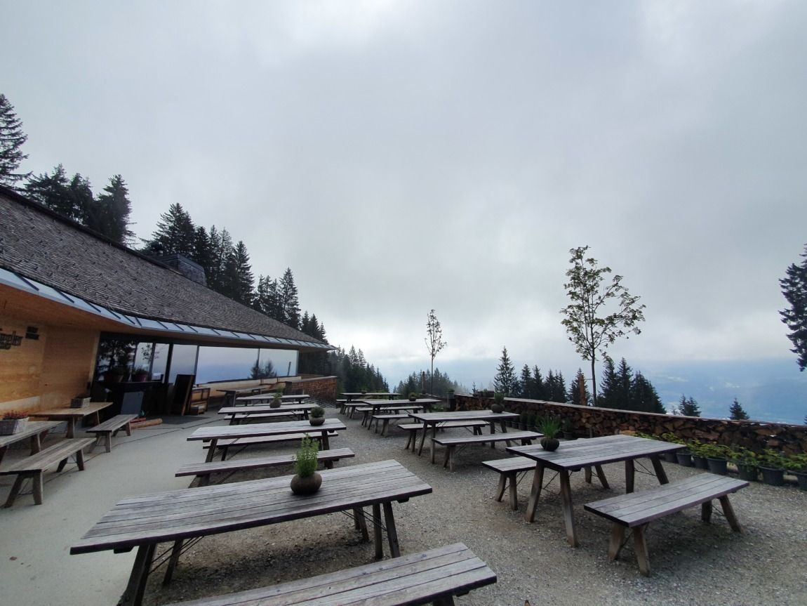 Freisitz mit leeren Picknicktischen aus Holz auf einer Terrasse in der Nähe der Umbrüggler Alm, umgeben von Bäumen und mit Blick auf eine wolkenverhangene Berglandschaft.