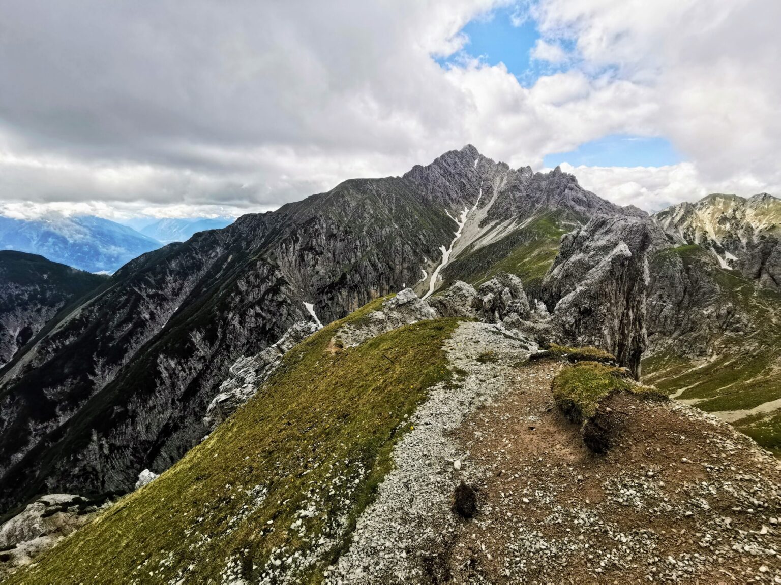 Ein schroffer Bergkamm mit felsigen Gipfeln und Grasflächen, wie geschaffen für eine Reither Spitze Bergtour, erstreckt sich unter einem teilweise bewölkten Himmel.