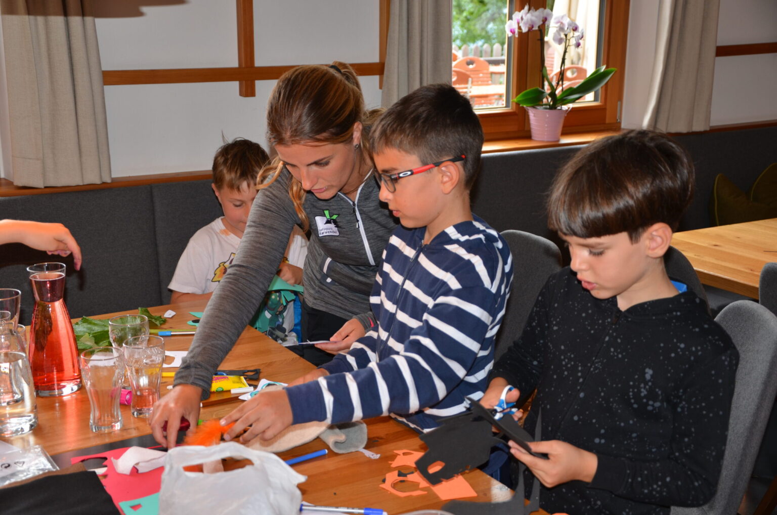 Eine Frau und drei Kinder sitzen an einem Tisch mit Bastelmaterial in einem gut beleuchteten Raum und genießen die Schulstunden, während sie Kunstprojekte erstellen, die vom Naturpark in der Schule inspiriert sind.