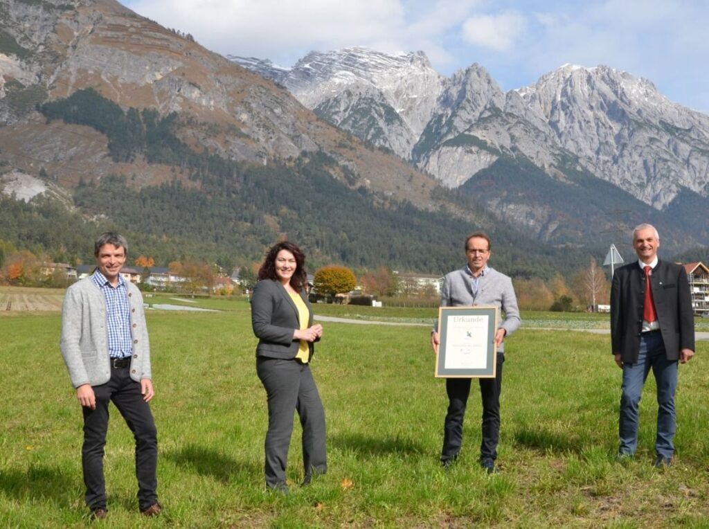 Vier Personen stehen auf einer Wiese im Naturpark Karwendel mit den Bergen im Hintergrund; eine von ihnen hält stolz eine gerahmte Urkunde.