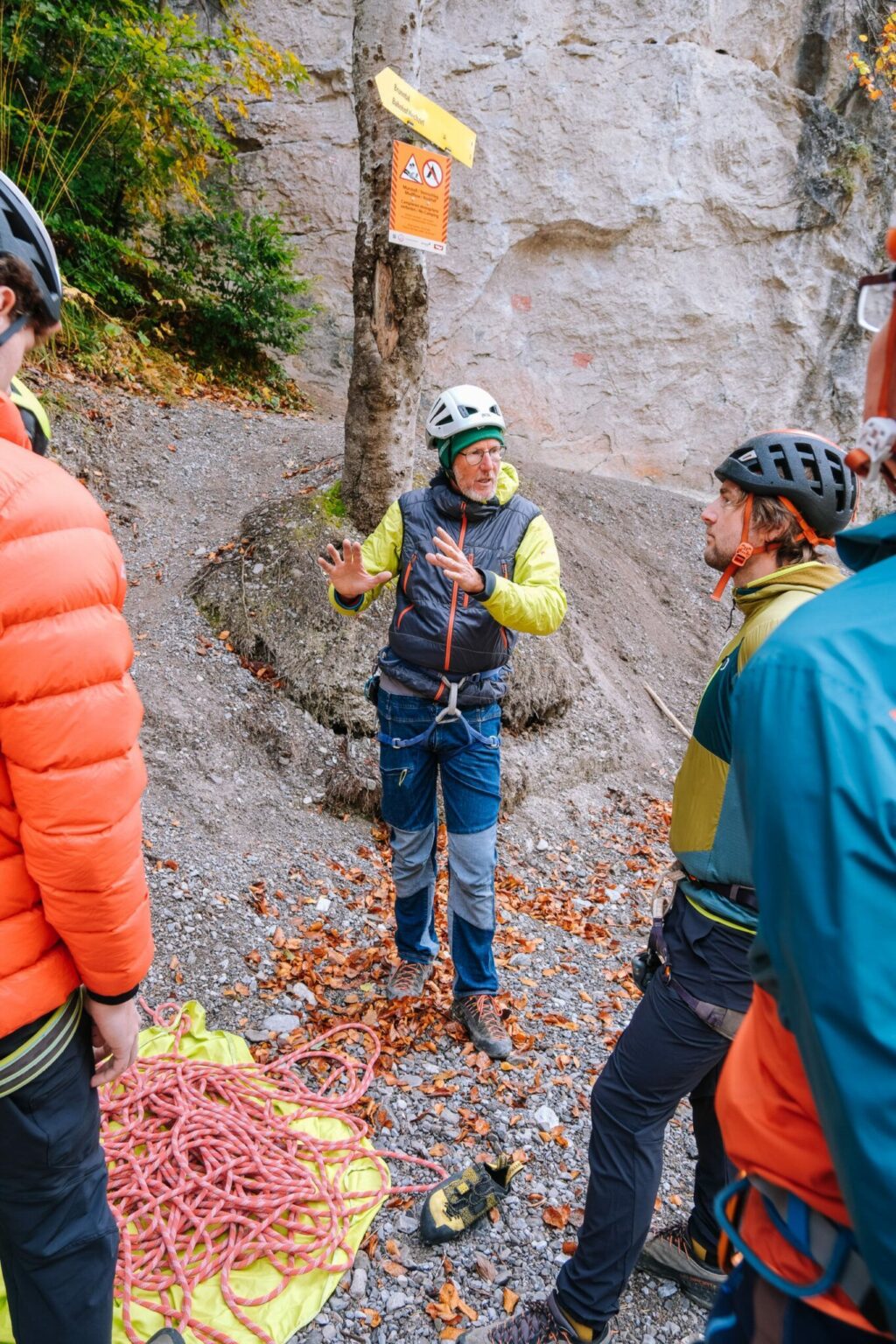 Eine Gruppe von Kletterern mit Helmen und Outdoor-Ausrüstung steht an einem aufgerollten Seil, während eine Person in der Nähe einer Felswand mit Warnschildern gestikuliert und spricht und betont, wie wichtig es ist, beim Klettern auf Sicherheit und Natur zu achten.