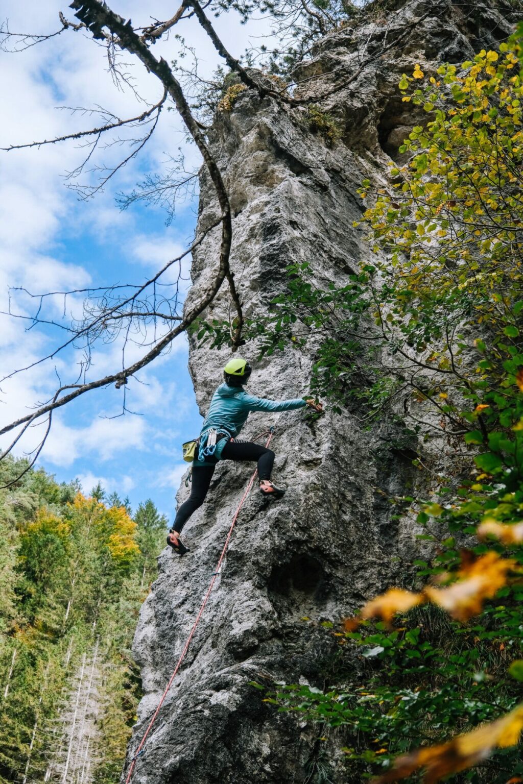 Eine Person, die einen Helm und eine Kletterausrüstung trägt, erklimmt eine steile Felswand in einem Klettercamp, umgeben von Bäumen und Grünflächen unter einem teilweise bewölkten Himmel.