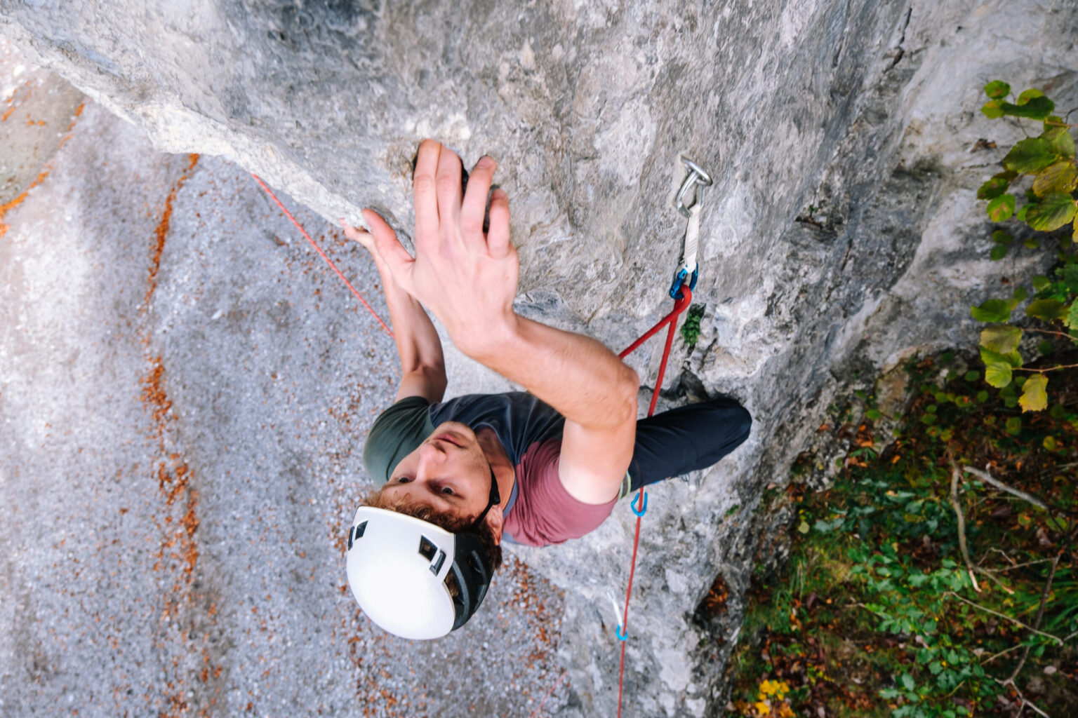 Eine Person, die einen Helm trägt, klettert an einem Sicherungsseil und einem Klettergurt eine senkrechte Felswand hinauf und demonstriert dabei ihre Kletterkünste, indem sie mit einer Hand einen Griff festhält und mit der anderen nach oben greift.