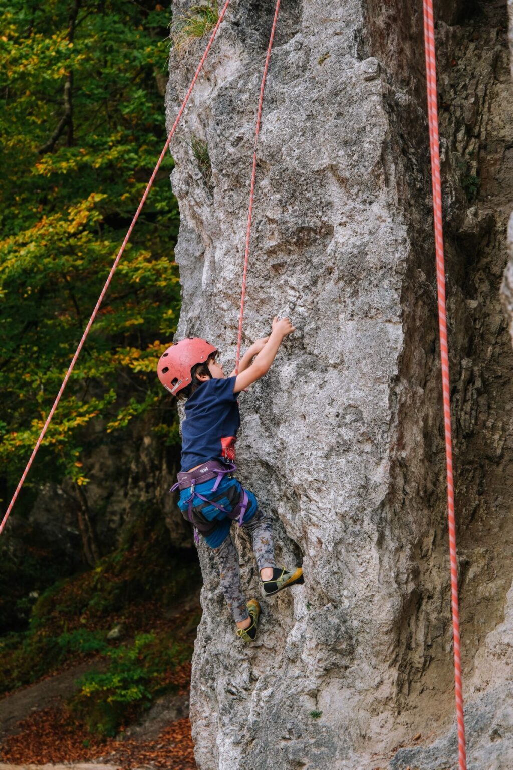 Ein Kind, das einen Helm und eine Kletterausrüstung trägt, klettert in der freien Natur an einem Seil eine Felswand hinauf, wobei es die Natur und die Sicherheit respektiert.