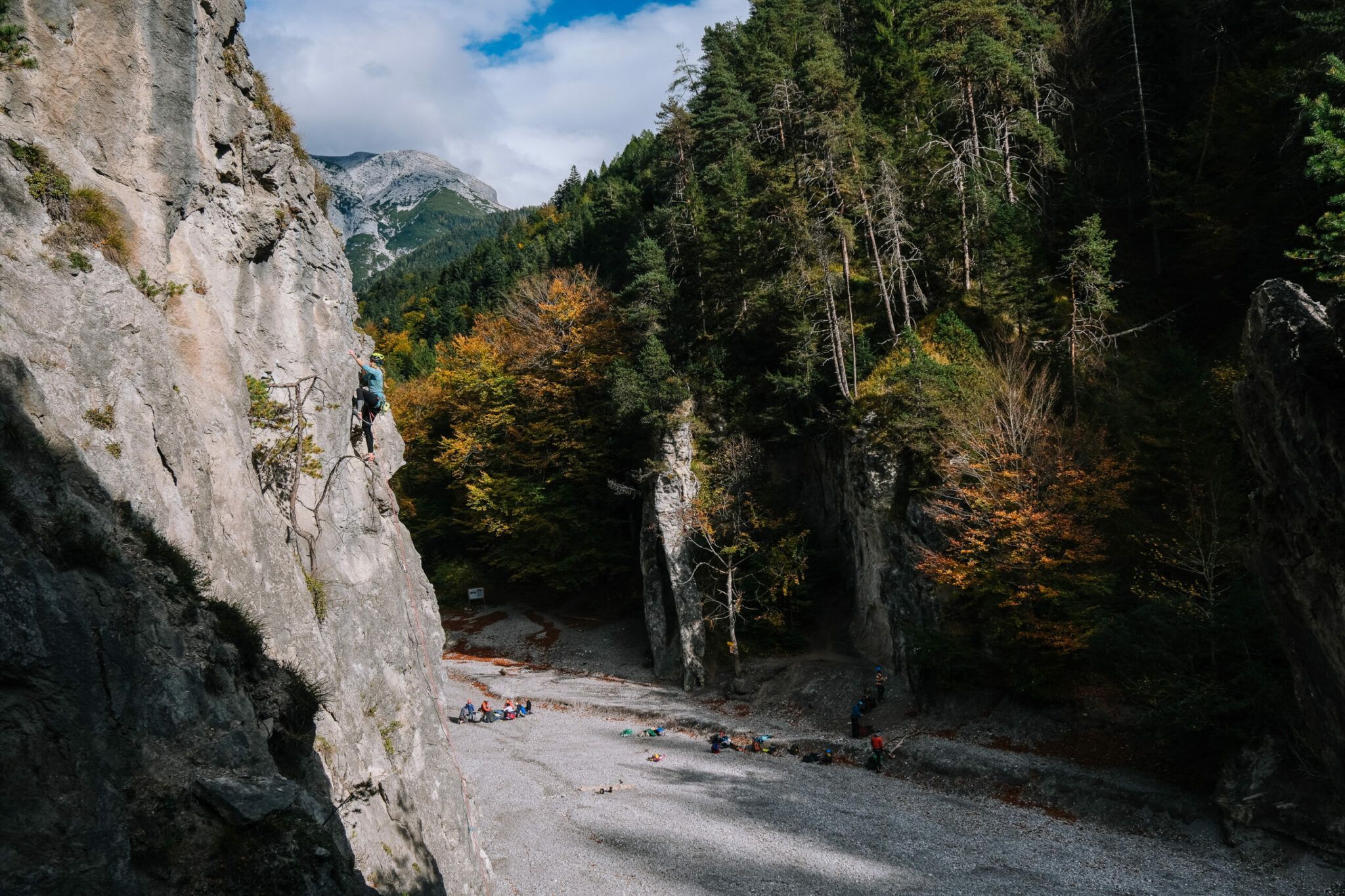 Eine Person klettert respektvoll eine vertikale Felswand auf der linken Seite hinauf und überblickt ein kiesiges Flussbett und die darunter versammelten Menschen, die alle von bewaldeten Bergen umgeben sind.