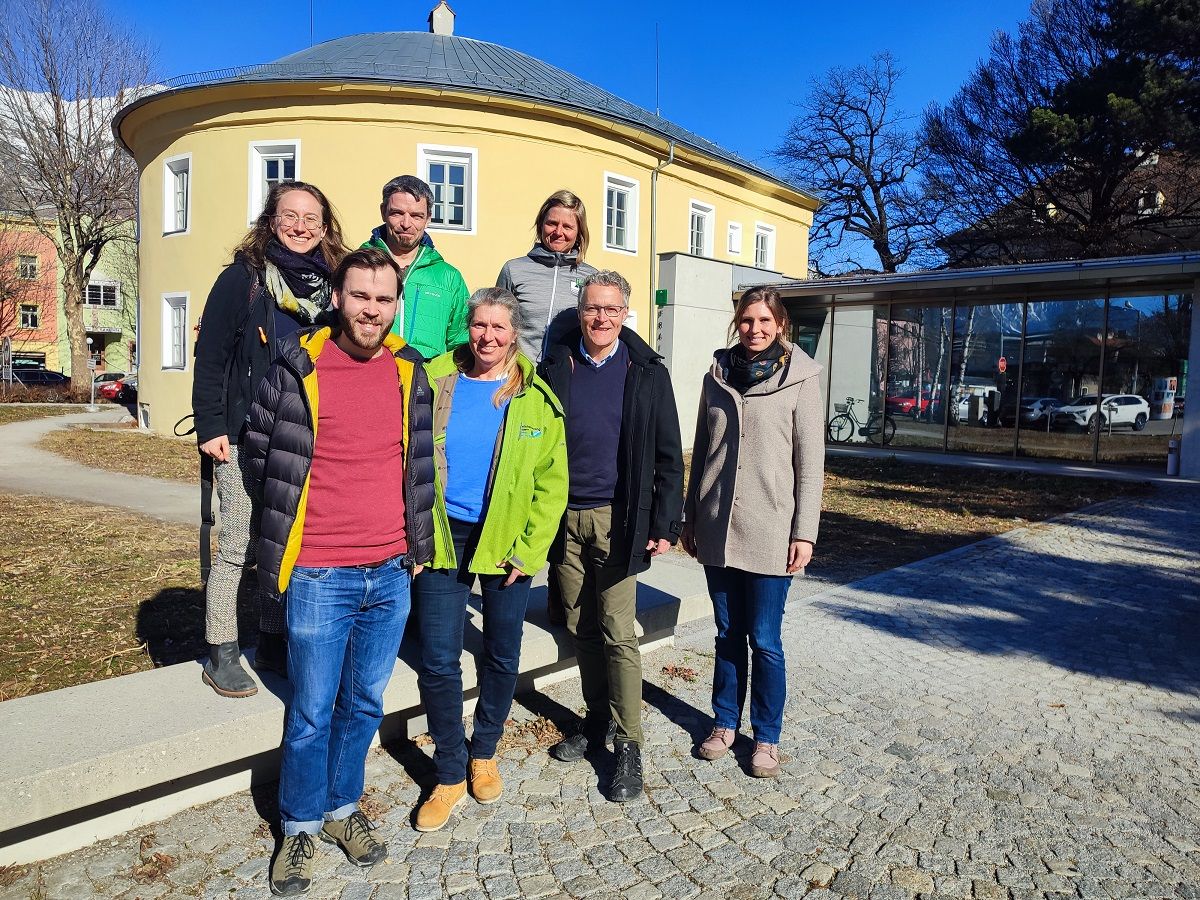 Sieben Erwachsene stehen an einem sonnigen Tag im Freien vor einem gelben Gebäude und posieren für ein Gruppenfoto auf einem kopfsteingepflasterten Weg - und fangen den Geist von AlmenREICH grenzenlos ein.