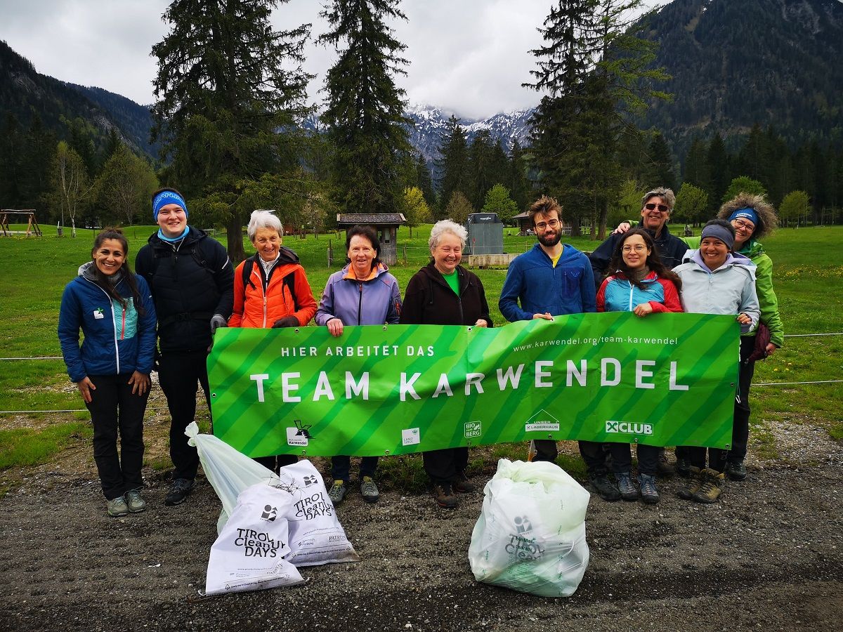 Eine Gruppe von Menschen, die im Freien ein grünes "Team Karwendel"-Transparent mit Säcken mit gesammeltem Müll in der Hand hält, nimmt an den CleanUp Days Karwendeltäler vor der Kulisse von Bergen und Bäumen am Achensee teil.