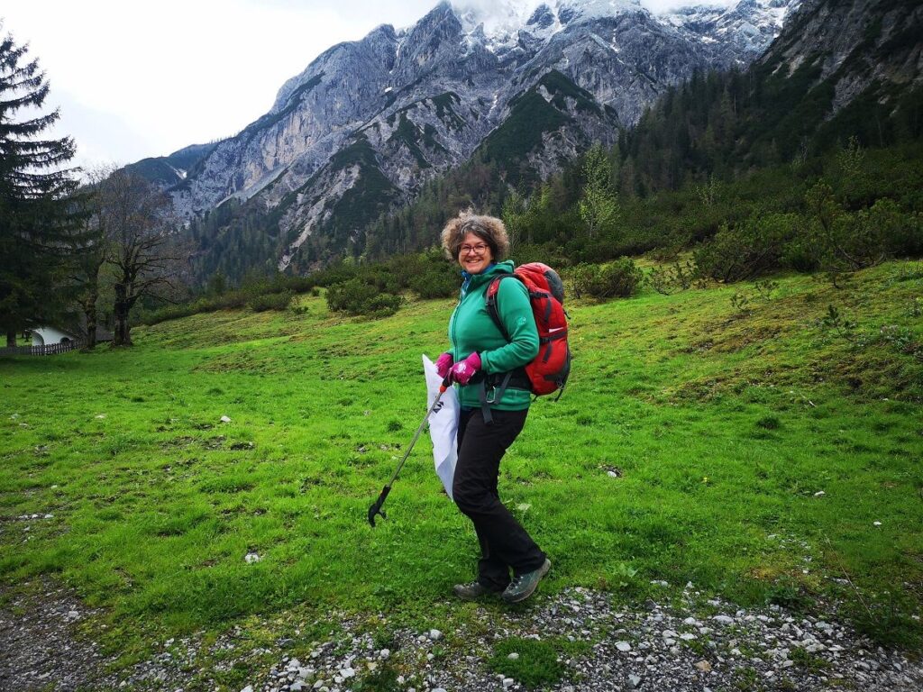 Eine Person mit rotem Rucksack und grüner Jacke steht während der CleanUp Days Karwendeltäler Achensee mit einem Wanderstock in der Hand auf einem grasbewachsenen Weg, mit Bergen und Bäumen im Hintergrund.