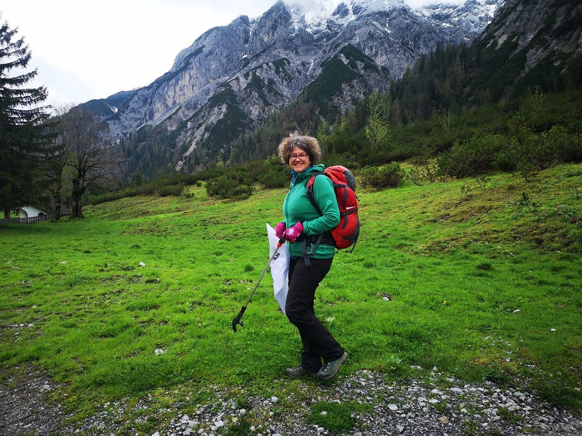 Eine Person mit rotem Rucksack und grüner Jacke steht während der CleanUp Days Karwendeltäler Achensee mit einem Wanderstock in der Hand auf einem grasbewachsenen Weg, mit Bergen und Bäumen im Hintergrund.