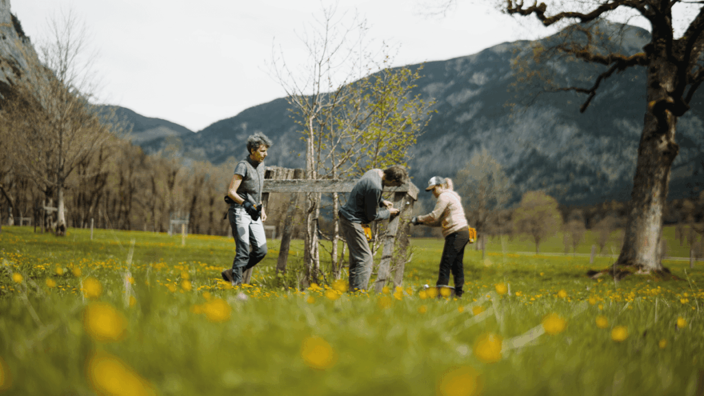 Drei Personen arbeiten im Freien an Zaunreparaturen auf dem Ahornboden und reparieren einen Holzzaun auf einer mit gelben Blumen übersäten Wiese, während sich im Hintergrund Berge und blattlose Bäume erheben.