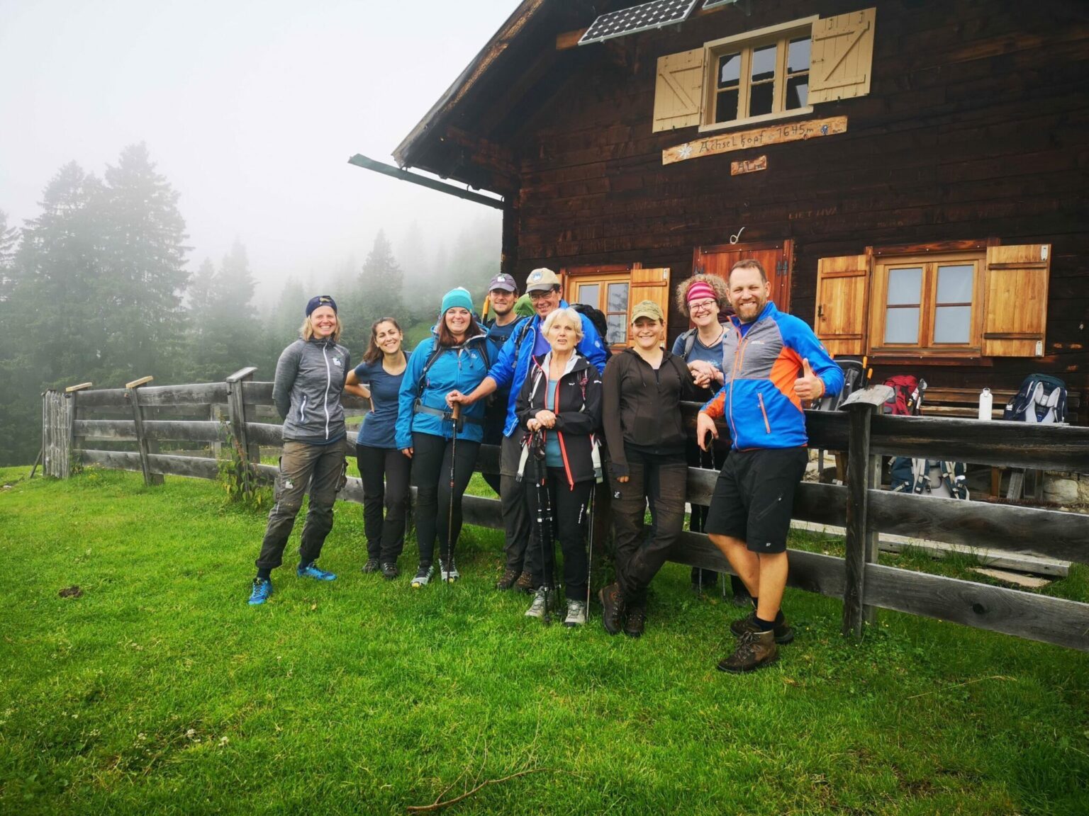 Eine Gruppe von Personen in Wanderkleidung steht und posiert gemeinsam vor der Almpflege Achselkopf Alm auf einer Wiese, mit nebligen Bäumen im Hintergrund.