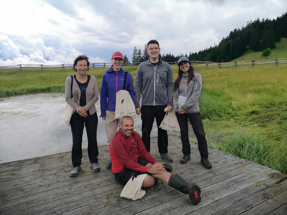 Fünf Personen in Outdoor-Kleidung posieren auf einer Holzplattform neben dem Walderalmteich in einer grasbewachsenen, hügeligen Gegend mit Bäumen und einem bewölkten Himmel im Hintergrund.