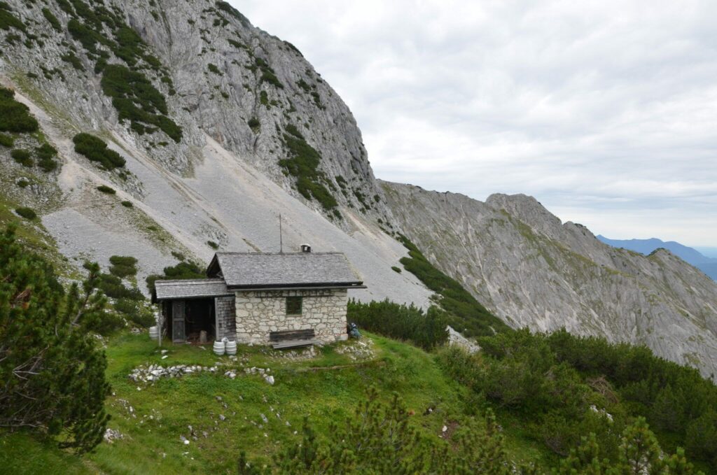 Eine kleine Steinhütte steht auf einem grasbewachsenen Hang, umgeben von Kiefernbüschen. Im Hintergrund erhebt sich steil das dramatische Arnspitzgebirge unter einem wolkenverhangenen Himmel.