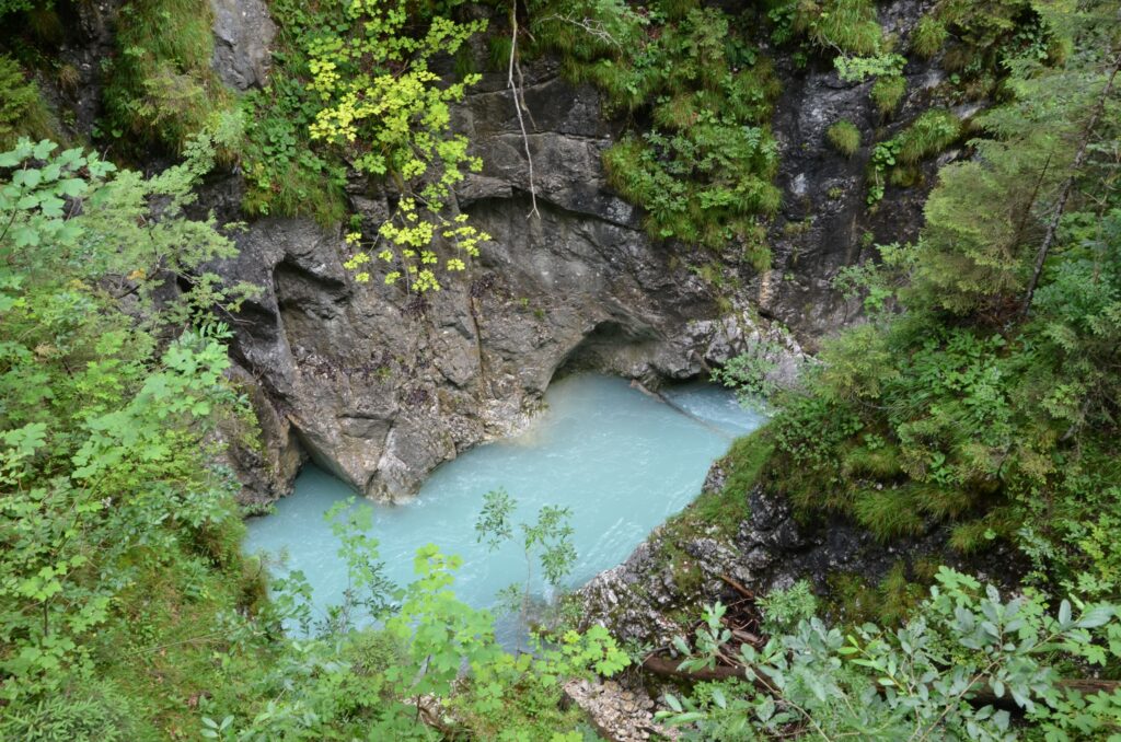 Ein kleiner türkisfarbener Bach fließt durch eine felsige Öffnung, umgeben von dichter grüner Vegetation und moosbewachsenen Steinen - eine einladende Szene auf der Wanderung Leutasch.