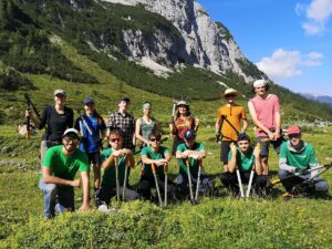Eine Gruppe von Menschen mit Werkzeugen posiert im Freien an der Umweltbaustelle Möslalm vor der Kulisse von Bergen und Grün unter einem klaren blauen Himmel.
