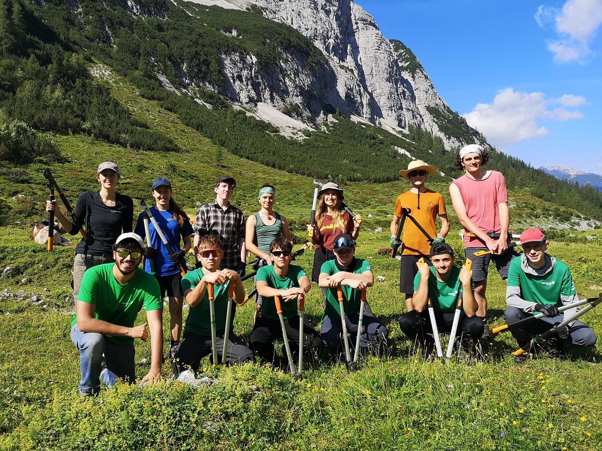 Eine Gruppe von Menschen mit Werkzeugen posiert im Freien an der Umweltbaustelle Möslalm vor der Kulisse von Bergen und Grün unter einem klaren blauen Himmel.