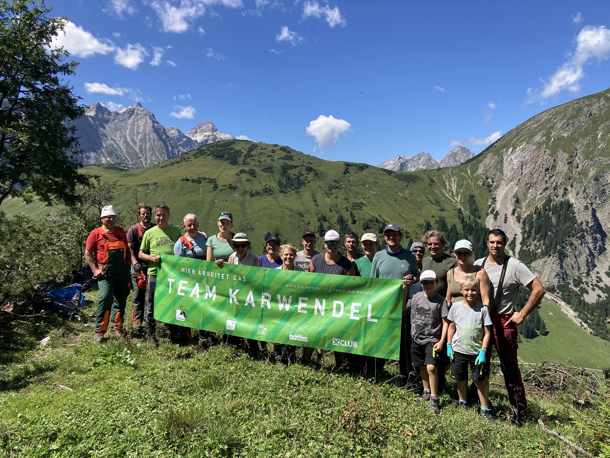 Eine Gruppe von Menschen in den Bergen, die bei strahlend blauem Himmel ein grünes "Team Karwendel"-Transparent halten und ihre Bemühungen um die Almpflege Lalidersalm feiern.