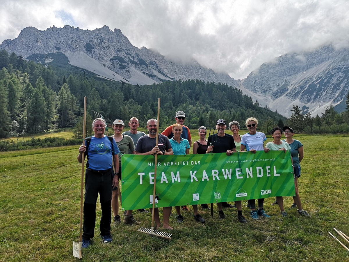 Eine Gruppe von Personen mit einem grünen "Team Karwendel"-Transparent steht im Freien auf einer Wiese mit Harken und nimmt an der Biotoppflege Issanger teil, mit Wald und Bergen im Hintergrund.