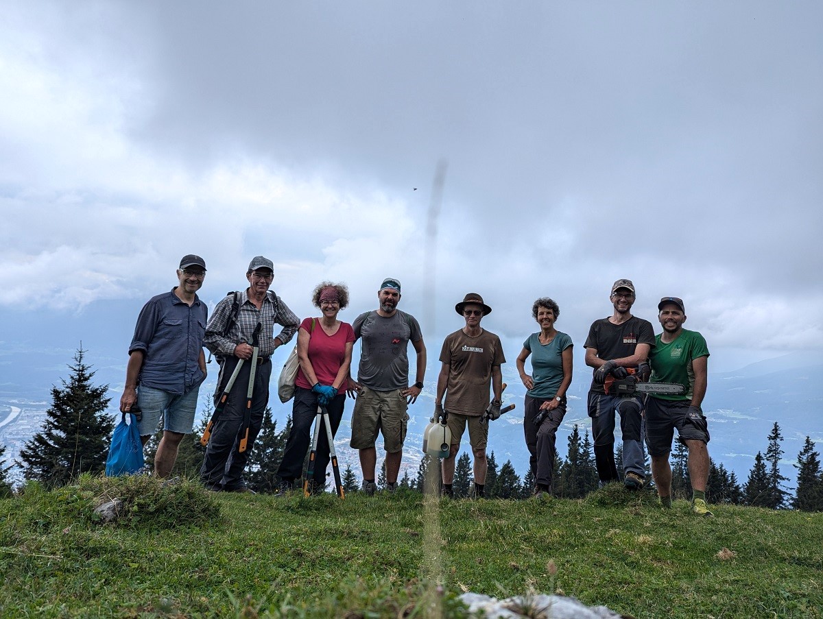 Eine Gruppe von acht Personen steht auf einem grasbewachsenen Hügel in der Nähe der Almpflege Achselkopfalm, mit Bäumen und bewölktem Himmel im Hintergrund, in der Hand eine Outdoor-Ausrüstung und lächelt in die Kamera.
