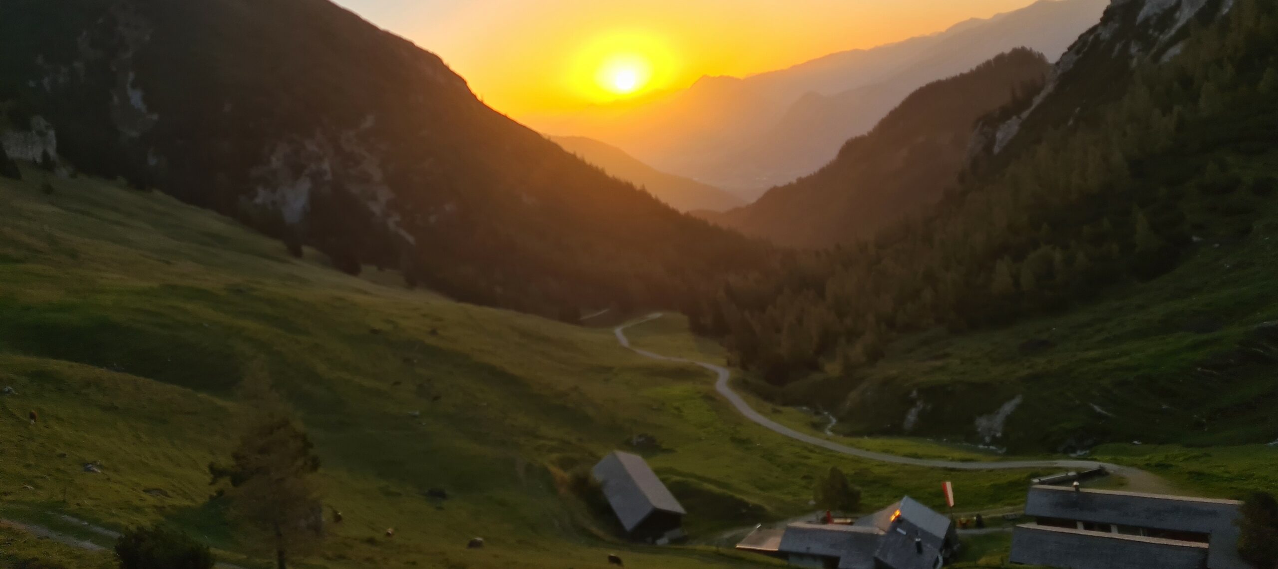 Sonnenuntergang über einem Bergtal mit einer kurvenreichen Straße, grasbewachsenen Hängen und ein paar Gebäuden im Vordergrund, die die ruhige Schönheit der Almpflege Weissenbachalm zeigen.
