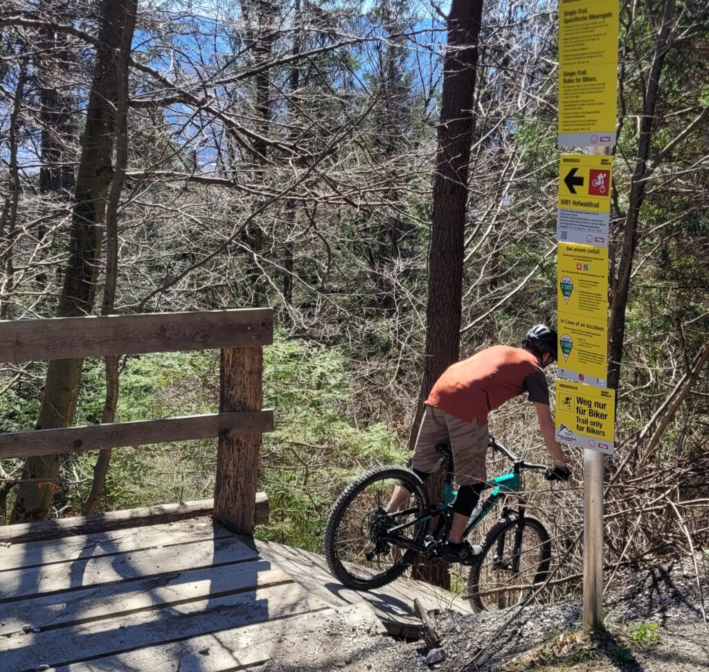 Ein Radfahrer, der einen Helm trägt, fährt mit seinem Mountainbike den Hofwaldtrail hinunter, vorbei an einem unbefestigten Weg in der Nähe einer Holzbrücke und einem hohen Wegweiser mit verschiedenen Wegmarkierungen in einem bewaldeten Gebiet.