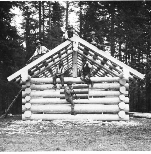Sechs Personen sitzen auf dem unvollendeten Holzgerüst einer Blockhütte im Hüttenstil in einem Waldgebiet, mit Bäumen im Hintergrund.