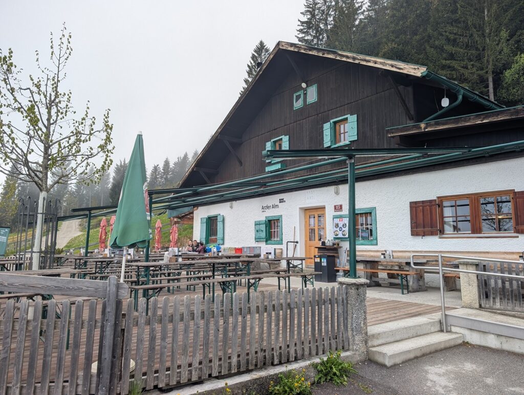 Ein rustikales Gebäude mit Sitzgelegenheiten im Freien, Holzzäunen und grünen Sonnenschirmen liegt an einem bewölkten Tag neben einem Waldgebiet und fängt den Charme der Almpflege Arzler Alm ein.