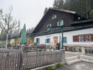 Ein rustikales Gebäude mit Sitzgelegenheiten im Freien, Holzzäunen und grünen Sonnenschirmen liegt an einem bewölkten Tag neben einem Waldgebiet und fängt den Charme der Almpflege Arzler Alm ein.