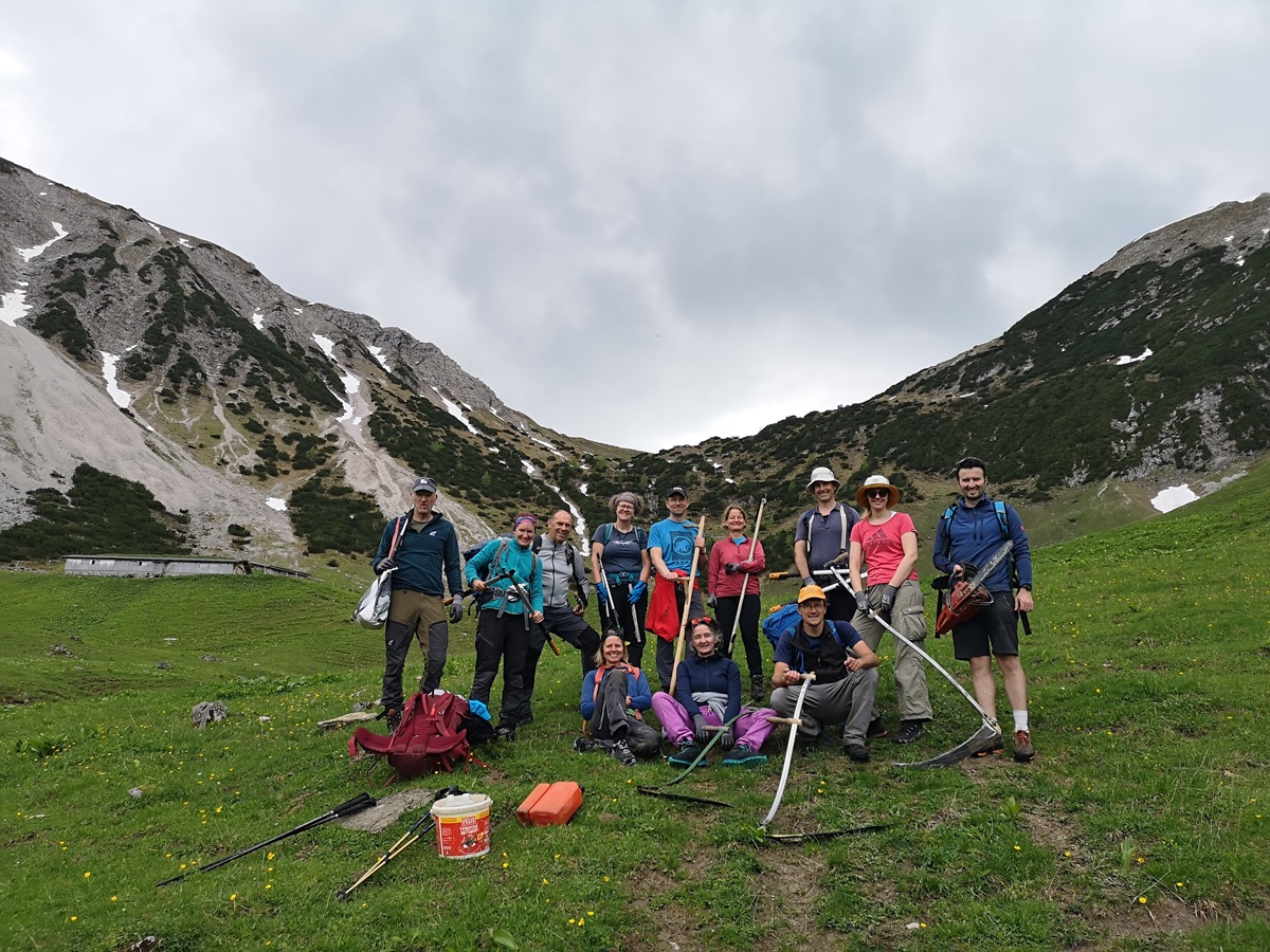 Eine Gruppe von Personen posiert im Freien auf einer grasbewachsenen Anhöhe der Almpflege Hölzelstalalm, mit Bergen im Hintergrund, und hält Werkzeuge und Ausrüstung unter einem bewölkten Himmel.