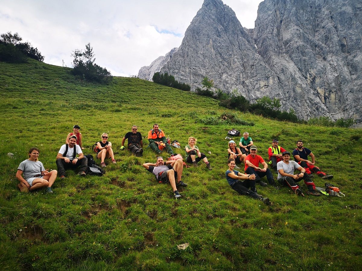 Eine Gruppe von Menschen sitzt auf einem grasbewachsenen Hügel bei der Almpflege Lalidersalm und entspannt sich, mit den Bergen im Hintergrund, an einem teilweise bewölkten Tag.