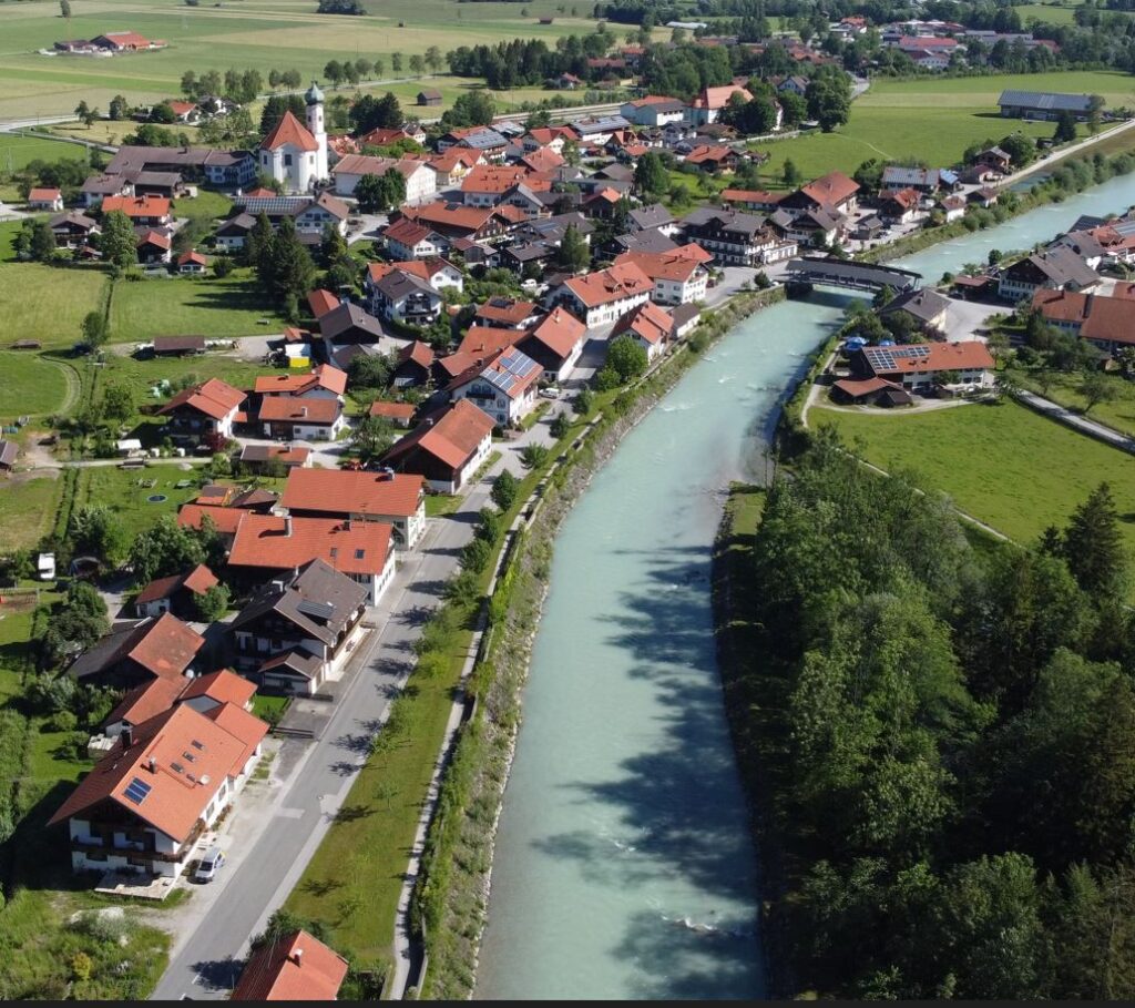Luftaufnahme eines kleinen europäischen Dorfes mit roten Dächern, einer Kirche und einem Fluss, der durch grüne Felder und Bäume fließt.