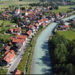 Luftaufnahme eines kleinen europäischen Dorfes mit roten Dächern, einer Kirche und einem Fluss, der durch grüne Felder und Bäume fließt.