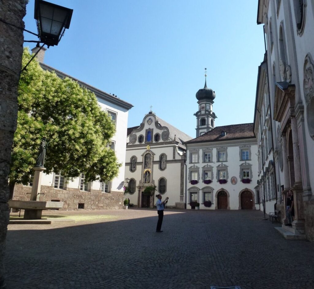 Eine Person steht in einem kopfsteingepflasterten Hof in Hall in Tirol, umgeben von historischen Gebäuden und einer Kirche mit einem Kuppelturm unter einem klaren Himmel.