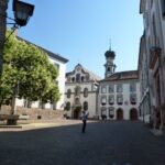 Eine Person steht in einem kopfsteingepflasterten Hof in Hall in Tirol, umgeben von historischen Gebäuden und einer Kirche mit einem Kuppelturm unter einem klaren Himmel.