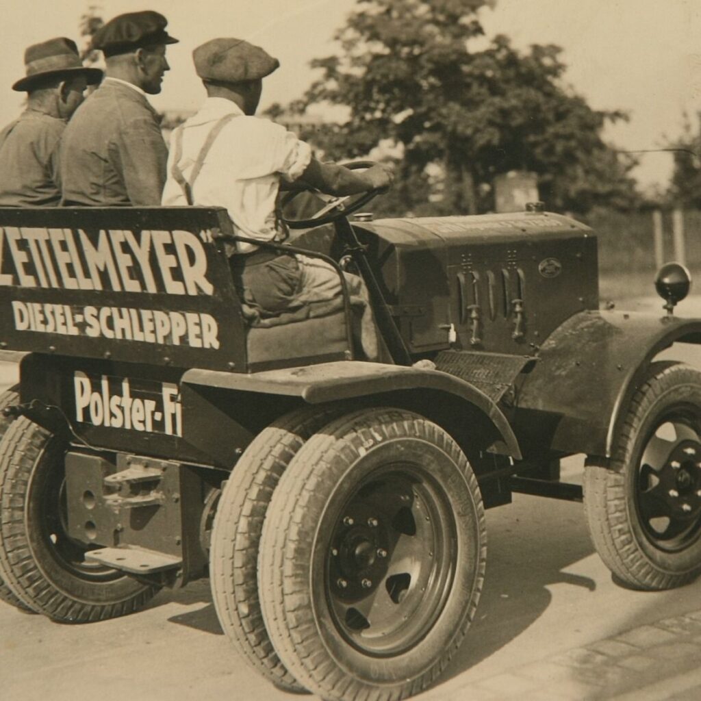 Vier Männer fahren auf einem alten Zetelmeyer-Dieseltraktor mit sichtbaren Werbeschildern auf der Rückseite, der auf einer asphaltierten Straße in der Nähe von Bäumen unterwegs ist.