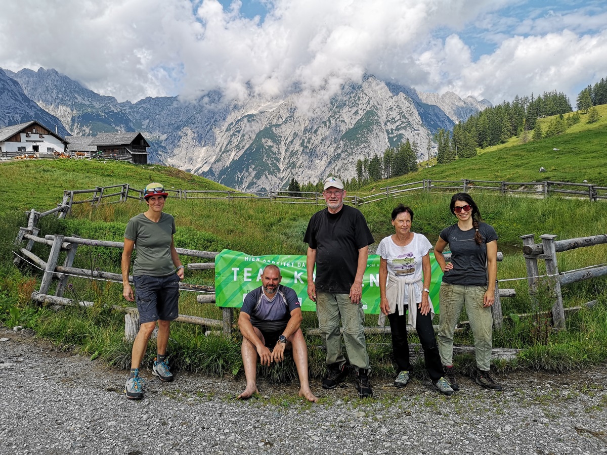 Fünf Personen posieren vor einem Holzzaun und einer grünen Fahne, während sich im Hintergrund die Berge, Wolken und grasbewachsenen Hügel in der Nähe der Walderalmteiche ausbreiten.