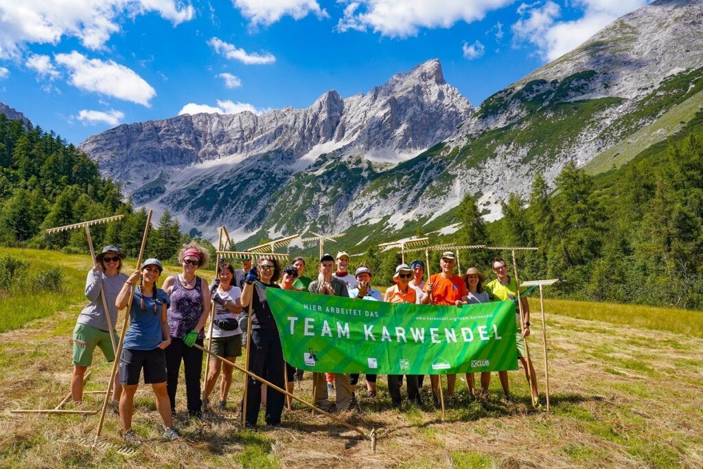 Eine Gruppe von Menschen steht mit Harken und einem grünen Banner mit der Aufschrift "Team Karwendel" auf der Issanger-Bergwiese, im Hintergrund felsige Gipfel und Bäume unter einem blauen Himmel.