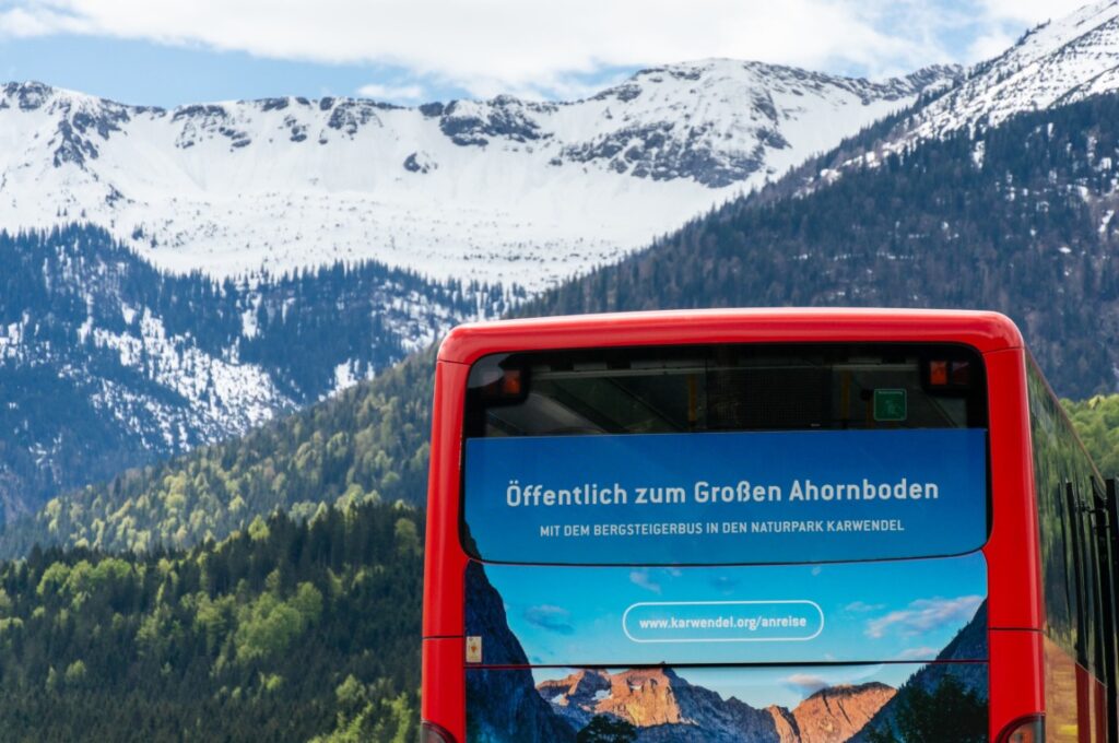 Ein roter Linienbus parkt vor den schneebedeckten Bergen und grünen Hügeln im Hintergrund. Der Bus zeigt ein deutsches Schild über den Service zum Großen Ahornboden im Naturpark Karwendel.